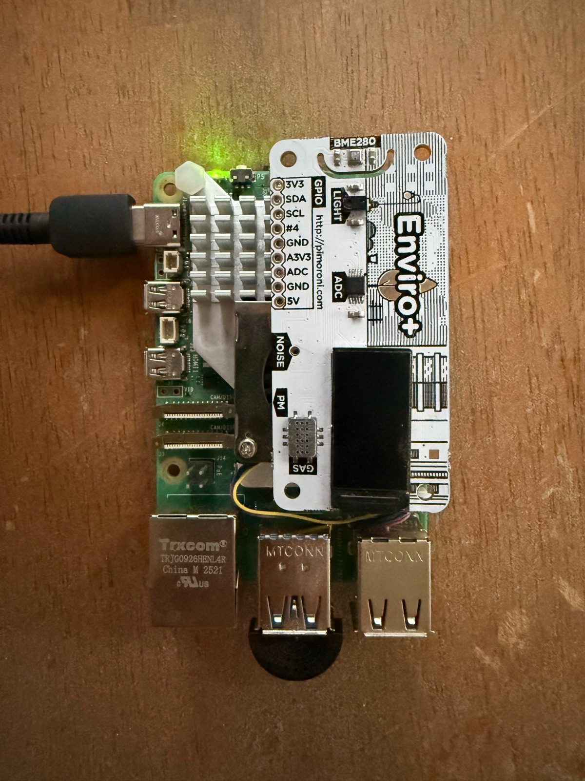 Raspberry Pi 5 on a wooden table with a Pimoroni Enviro+ board mounted on top and a small black USB microphone in one of the USB ports